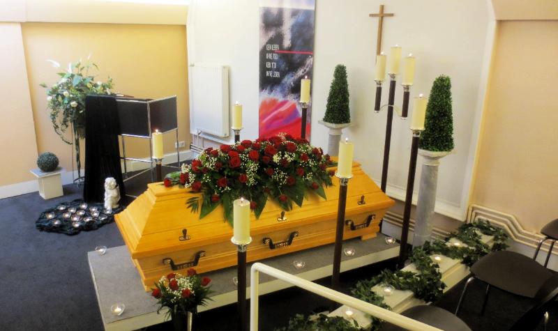 Bild eines Sarges, der geschmückt ist und in einer Trauerhalle steht. Kerzen stehen rundherum.